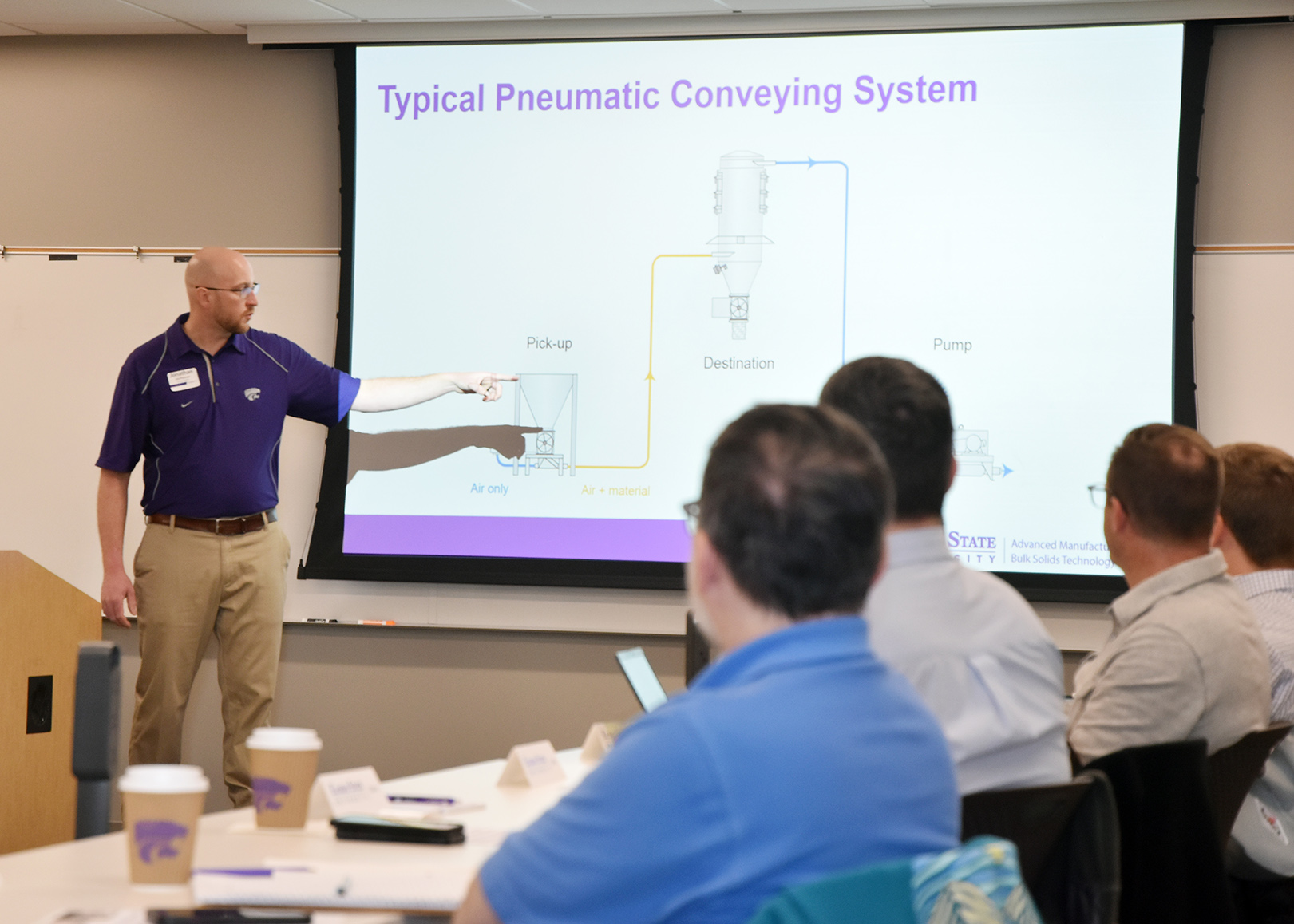 An instructor in a purple polo shirt points to a Powerpoint presentation in front of a class of students.
