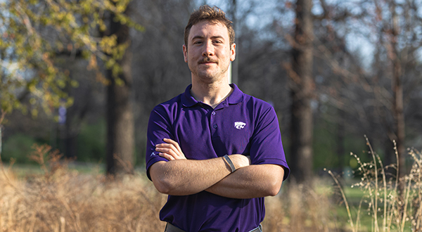 A male college student in a purple polo poses for an outdoor portrait, with wooded prairie areas behind him.
