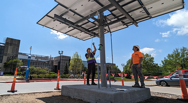 Two people in hardhats stand under a large solar panel outside with blue skies in the background