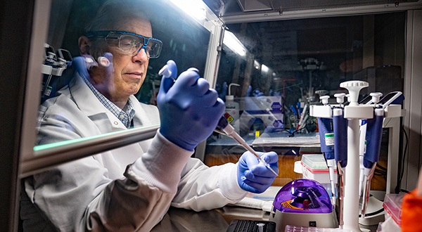 A man in a white lab coat and blue gloves with safety glasses works with tubes in a lab behind a protective hood
