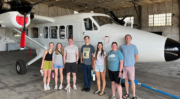 A college researcher and students in his lab pose for a group photo in front of an airplane in a hangar.