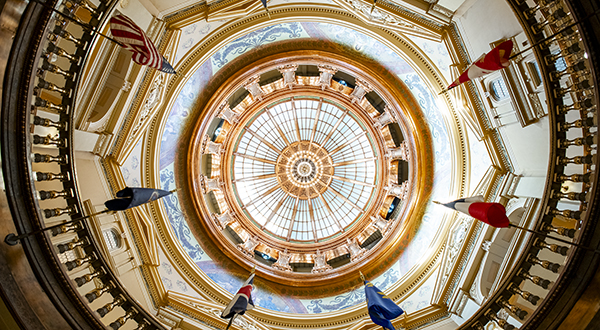 A bottom view of the Kansas Statehouse rotunda shows its impressive circular archiecture, with a ring closest to the camera showing the second floor balcony and several country's flags posted off the balcony railings.