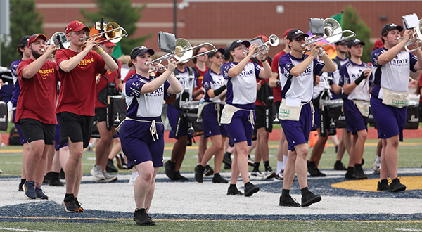 Alternately wearing purple-and-white and red uniforms, members of two separate marching bands rehearse jointly on a football field.