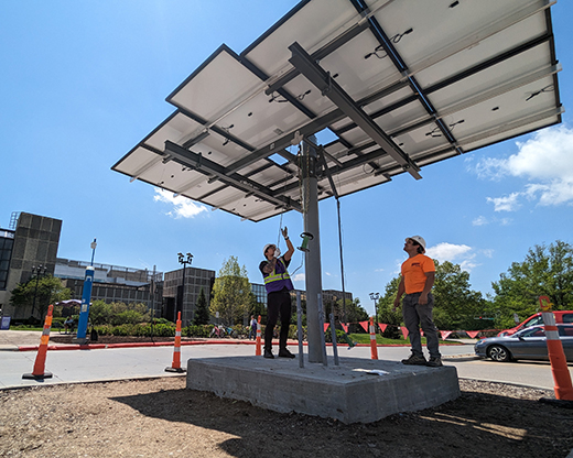 A student in a purple safety vest pulls on a hoist system that gradually pulls a large array of solar panels up a metal column.