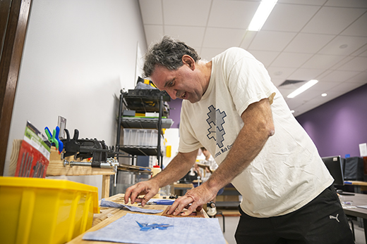 Wearing a white shirt with a puzzle of shapes and the words "Indigenous Math Circles," Dave Auckley leans over a wooden counter topped with various tools and items as he cuts apart the plastic shapes to make his math puzzles.