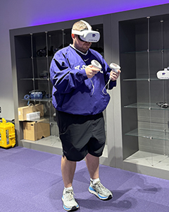A student in a purple hoodie stands and uses a white virtual reality headset, as well as handheld paddles used to manipulate the virtual environment.
