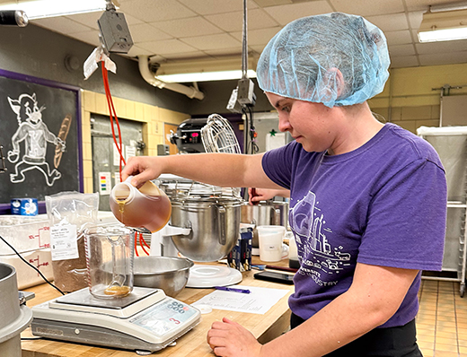 Bakery science student pouring honey A student in a purple shirt and sanitary cap pours honey into a measuring cup