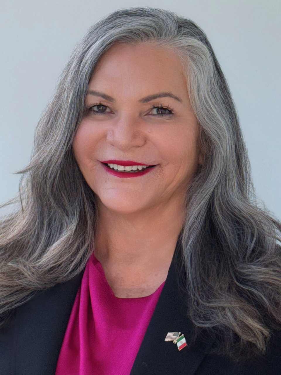 A woman with dark and gray hair smiles against a light gray backdrop. She is wearing a black blazer over a pink.