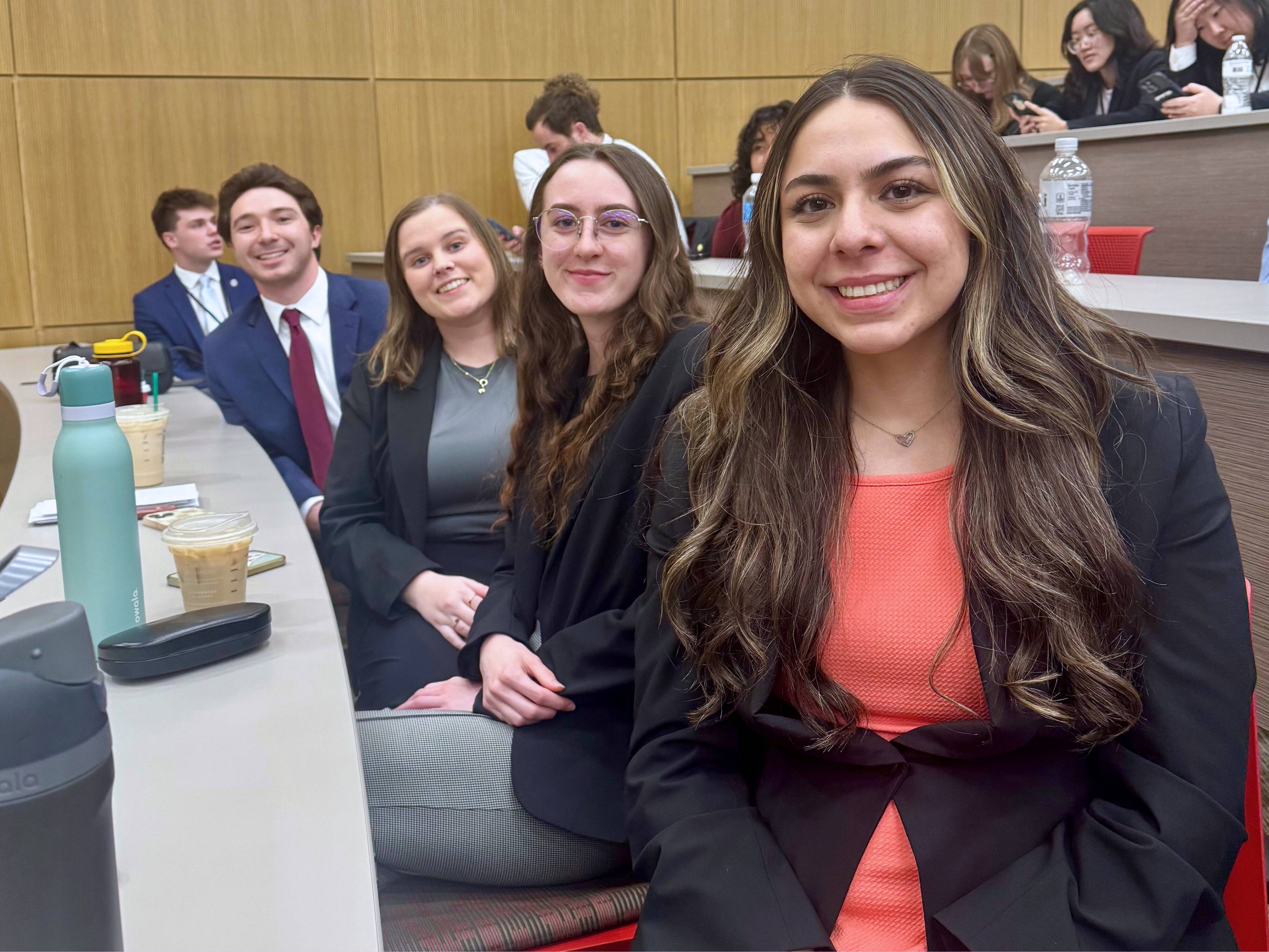 Four business students in professional attire sit at a long table in a lecture-style classroom.