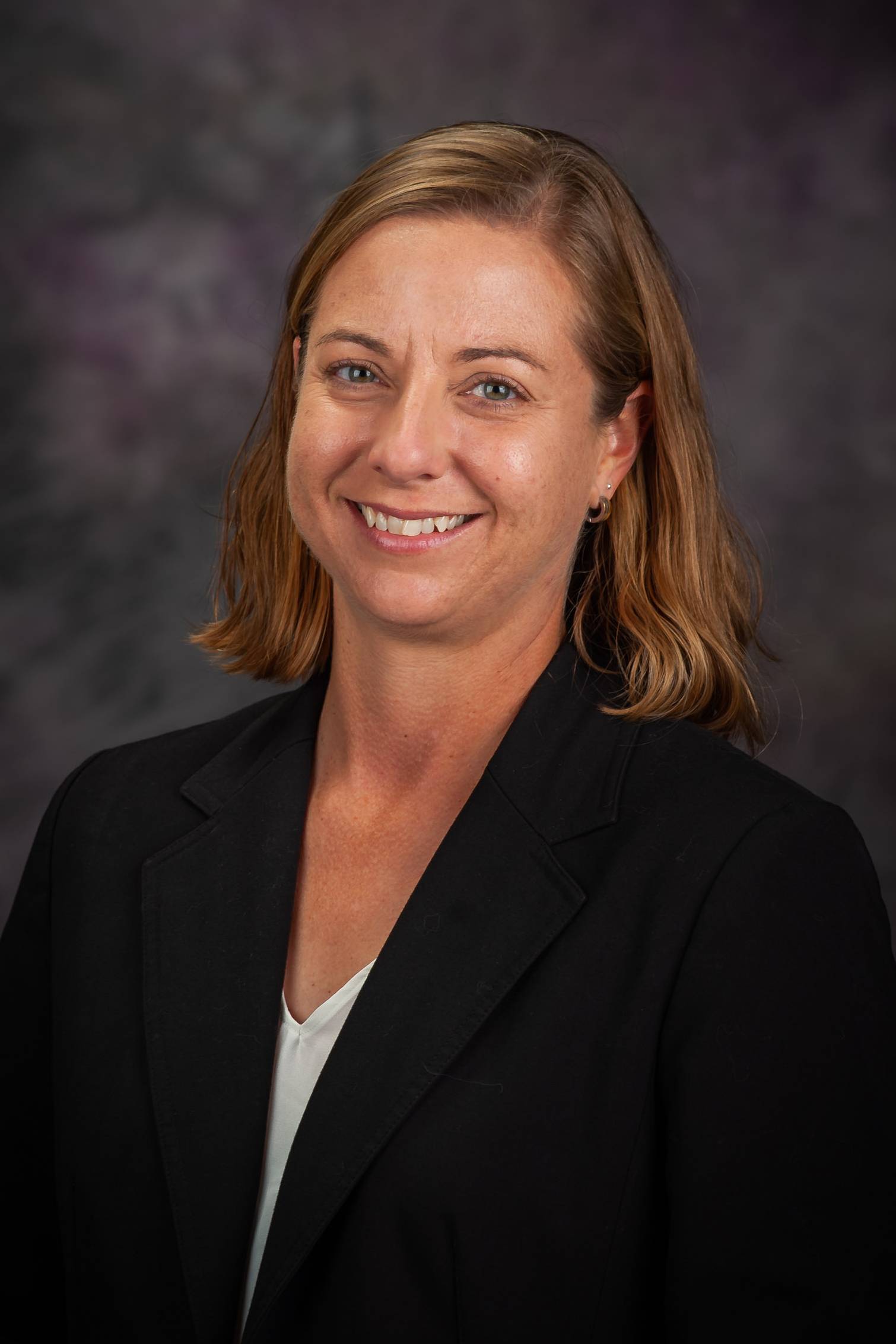 A woman with short, light-brown hair wearing a black blazer over a white shirt smiles for a portrait photo against a dark gray backdrop.