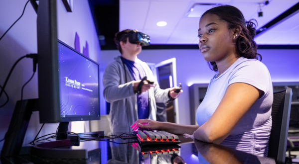 A student works on a computer while another student uses VR.