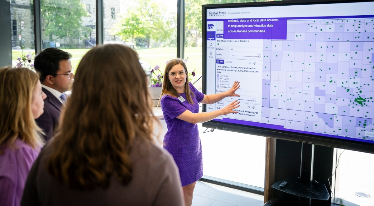 A woman in a purple dress presents how to use an interactive map software on a TV to a group of people