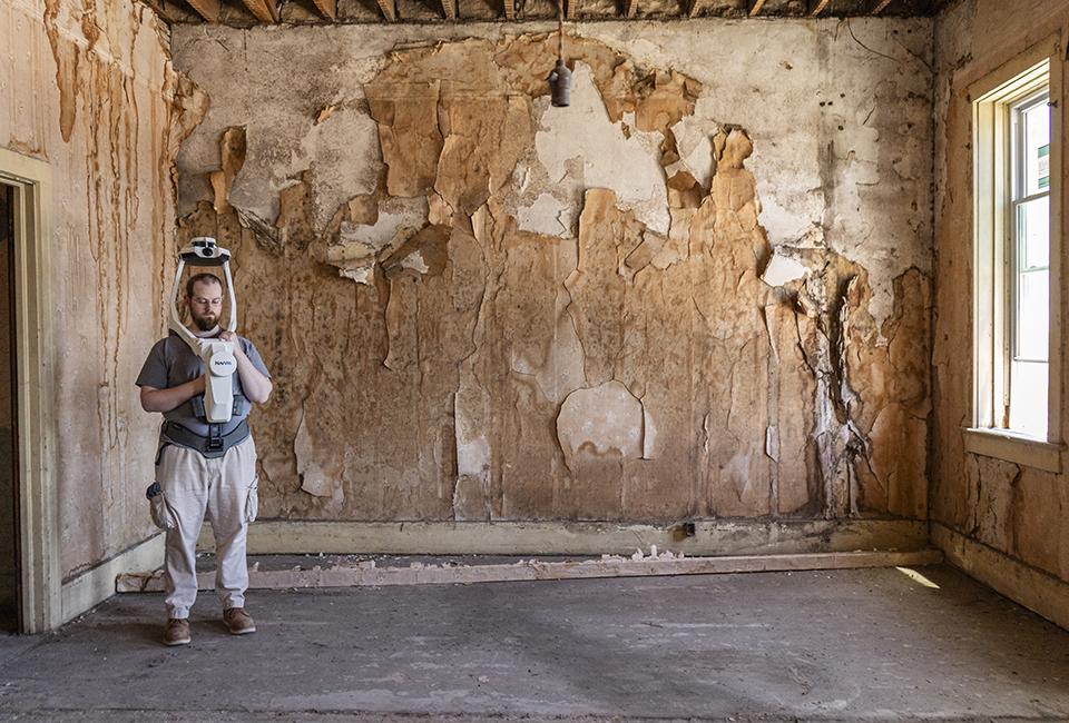 A man wearing a shoulder-mounted, above-head camera system stands in an empty room in front of a wall with peeling wallpaper.