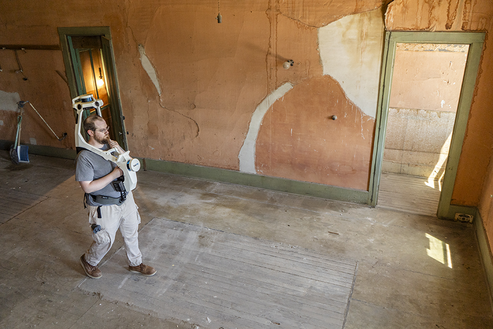 A man, wearing a shoulder-mounted, overhead 3D camera, walks across an empty room as he uses the camera to scan and pinpoint a 3D map of the area.