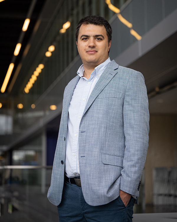 A researcher in a gray blazer poses in front of a glass barrier in a dimly lit academic building's atrium.