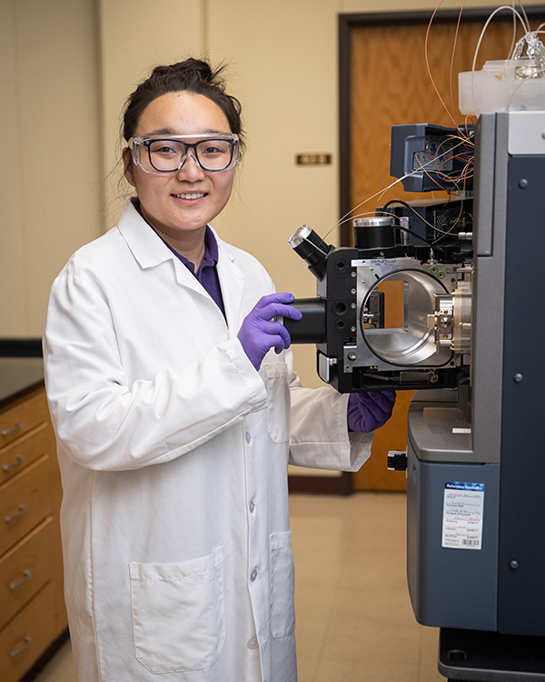 A researcher in a white lab coat and purple gloves near machinery.