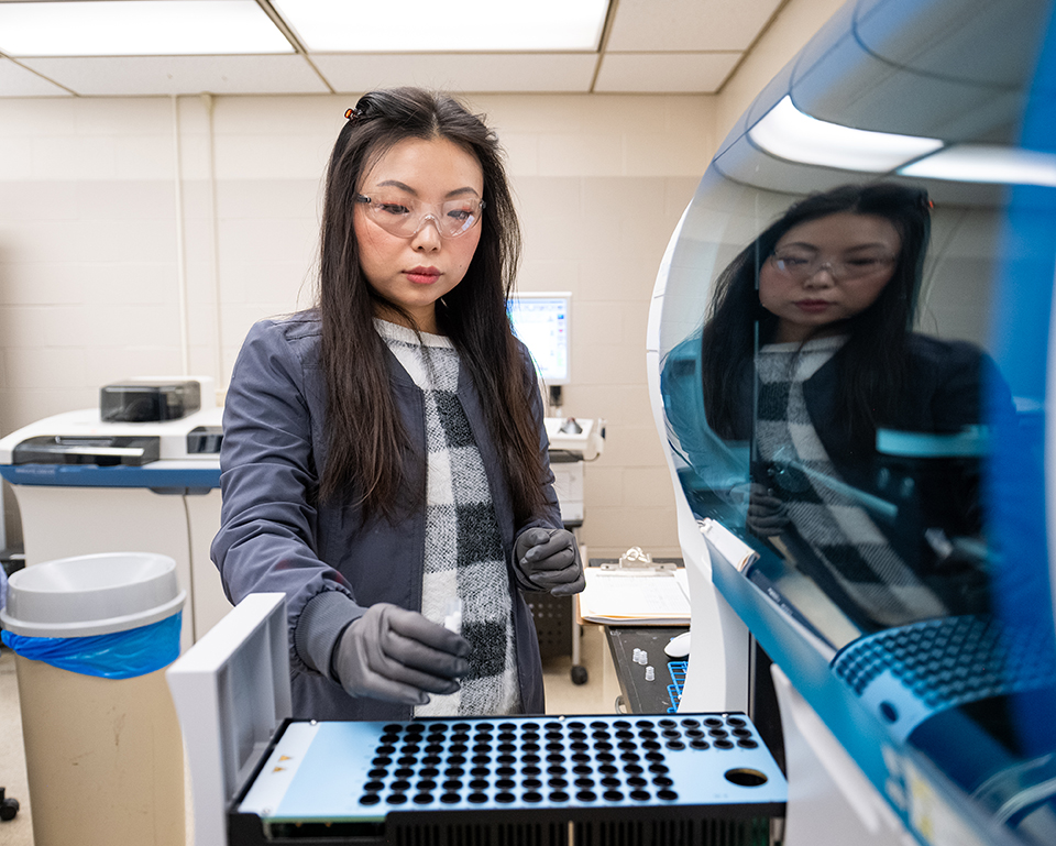 A woman wearing safety goggles loads a blood sample into one of several holes in a tray sticking out of a blue blood clotting machine.