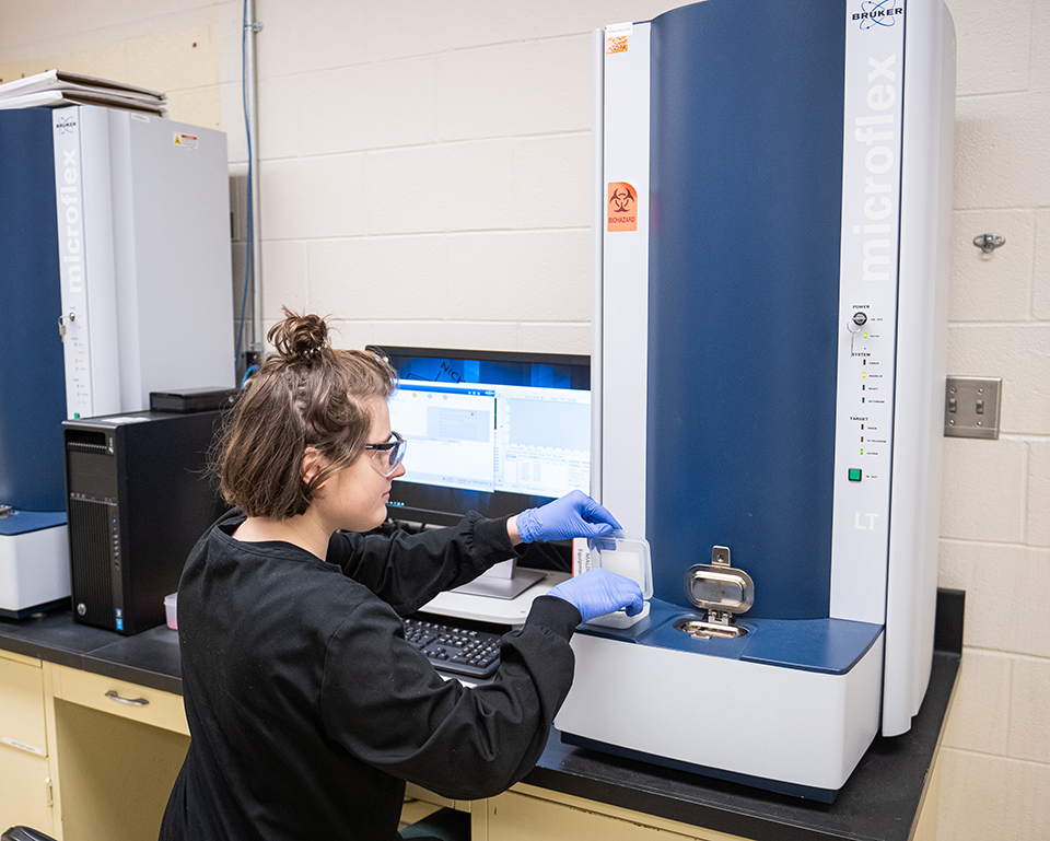 Facing a wall, a woman loads a target into a tall, mass spectrometer used to identift microorganisms.