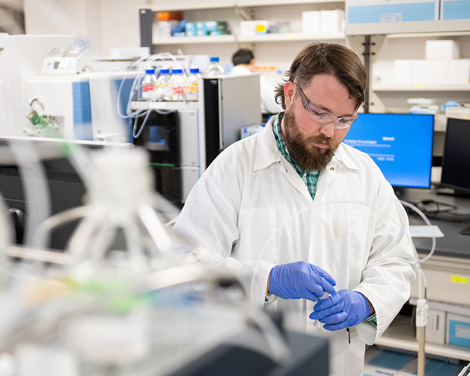 A man wearing safety goggles and blue rubber gloves loads a sample into a plasma mass spectrometer inside a veterinary testing laboratory.