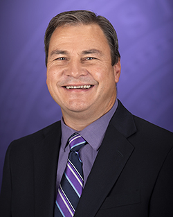 A man in a dark suit with a purple shirt and tie smiles ahead for a portrait.