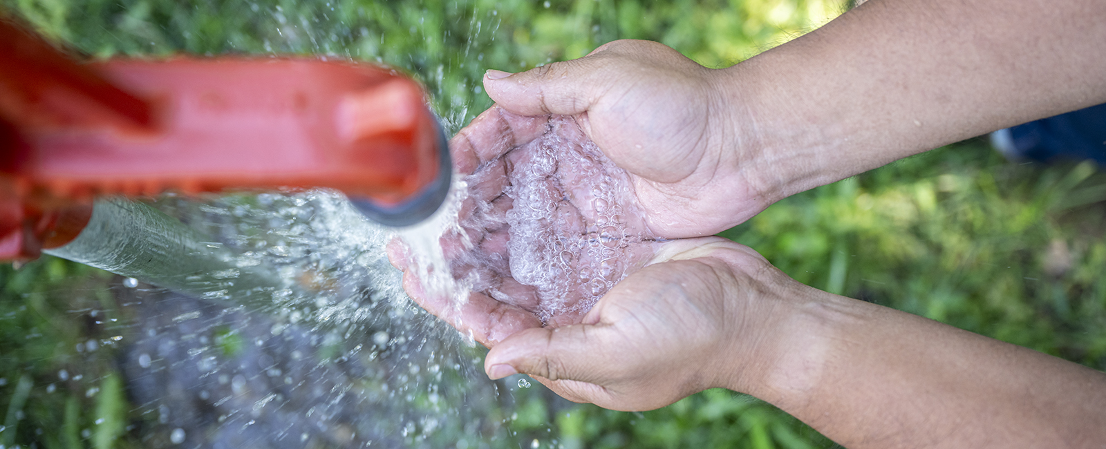 A topdown view of a pair of hands reaching in from the right side, palms up as water flows down from a red spigot onto the hands.