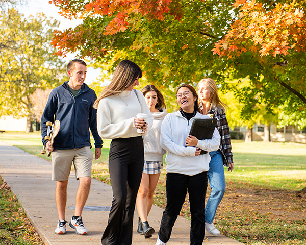 A group of students walks on a sidewalk during a sunny fall day, with brightly colored trees behind them.