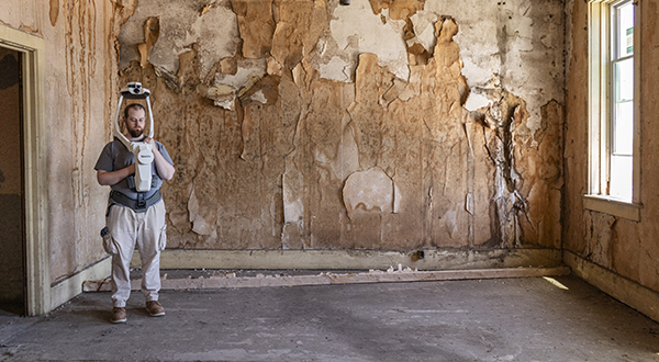 A man with a shoulder-mounted, above head camera scans a room with peeling wallpaper.