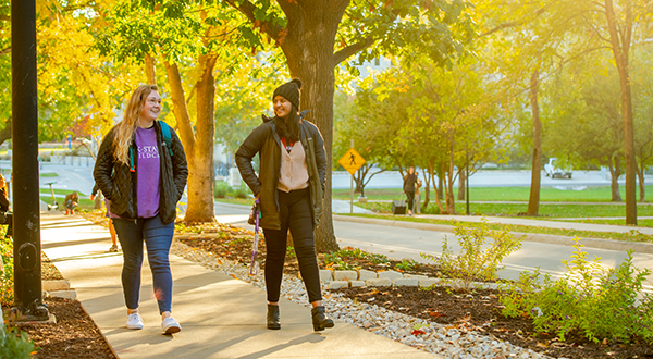 A pair of students walk up a sidewalk as brilliant beams of sunlight poke through surrounding trees.