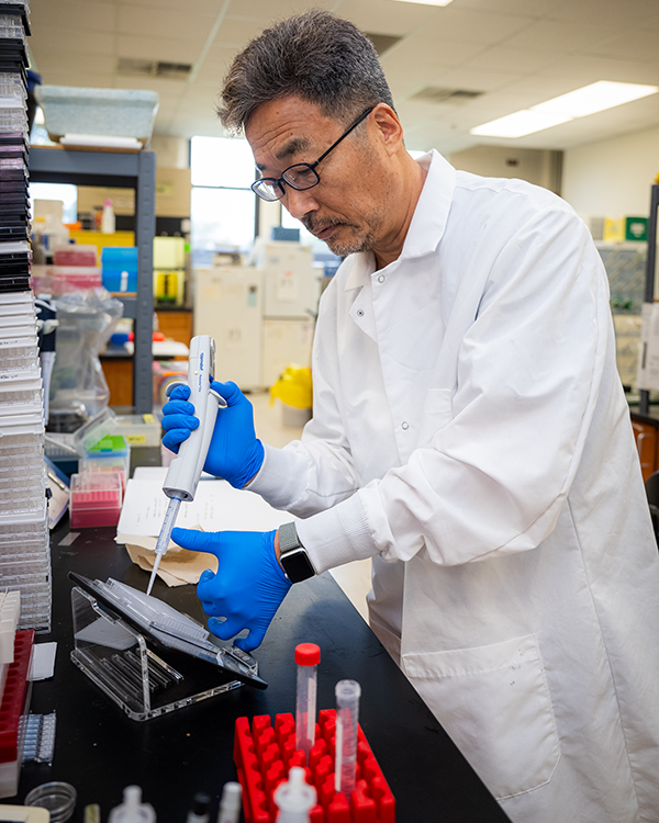 A man in a white lab coat and glasses uses a pipette on a work bench in a laboratory.