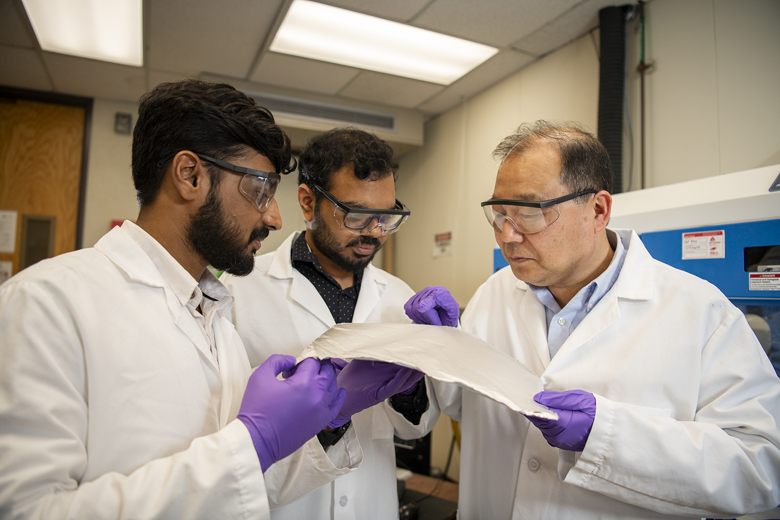 A group of researchers wearing white lab coats and purple gloves hold a sheet of 3D nanoprinted material.