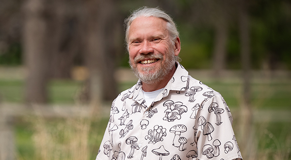 A man in a shirt with several musrhooms printed on it smiles for an outdoor portrait.