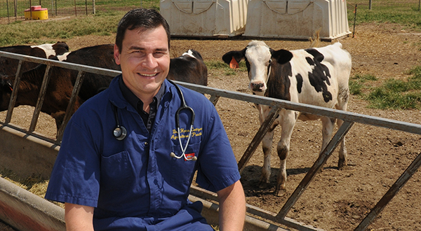 A man in a blue work coat poses for a portrait in front of a cattle trough, with a black-and-white spotted cow behind him.