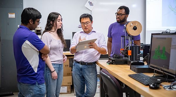 In an engineering lab, a professor holds up a tablet computer and explains concepts to a group of three undergraduate students who listen around him.